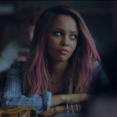 a woman with pink hair sitting at a table in front of other people and looking off into the distance