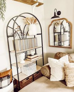 An arching iron and wood bookshelf is given a farmhouse style with neutral woven storage baskets, book covers, and white stone vases and figurines. The shelf is placed along the side of a taupe sofa with neutral throw pillows and stands before a wood-framed mirror and a black sconce light...   Image: our.nohai.home