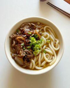 a white bowl filled with noodles and meat on top of a table next to chopsticks