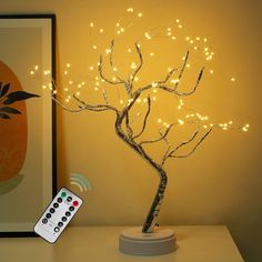 a lighted tree with remote control sitting on top of a table next to a painting