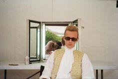 a man sitting in front of a mirror with sunglasses on his face and wearing a vest