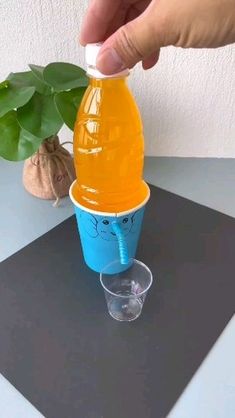 a hand is holding a plastic cup with an orange liquid in it and a plant next to it