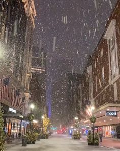 a snowy street with buildings and flags in the background at night time, as snow falls on the ground