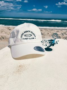 "Rep one of our favorite beach towns with this trendy hat!  This hat style features our Surf Club graphic and a low profile with an adjustable strap and curved visor. Perfect for those sunny beach days! This embroidered black and pink design comes in: Pink, Light Blue, Stone, Khaki, and White. Item Features: * 100% chino cotton twill * Unstructured, 6-panel, low-profile * 6 embroidered eyelets * 3 ⅛\" (7.6 cm) crown * Adjustable strap with antique buckle *Please note: Color may differ slightly from how it appears on your screen due to varying monitor settings.  Some designs may come with a white paper backing on the inside of the embroidered design, this is needed during the creation process to ensure the strength and longevity of the design. This product is made especially for you as soon Cheap Pink Baseball Cap For Beach, Vacation Bucket Hat With Curved Brim, Beachy White Hats For Poolside, Curved Brim Bucket Hat For Vacation, White Snapback Baseball Cap For Travel, White Snapback Hat For Travel, White Sun Hat For Vacation Poolside, White Hat For Poolside Vacation, White Sun Hat For Poolside Vacation