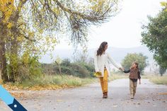 a woman holding the hand of a little boy walking down a road with trees in the background