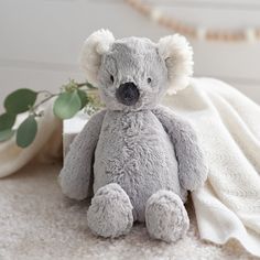 a gray teddy bear sitting on top of a white blanket next to a green plant