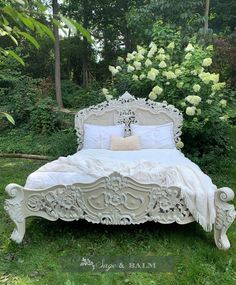 a white bed sitting in the middle of a lush green field next to trees and flowers