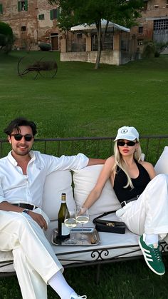 a man and woman sitting on a white couch in the grass with wine bottles and glasses