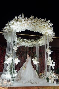 a woman in a wedding dress standing under a white flower covered arch with candles and flowers