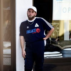 a man standing in front of a window with his hands on his hips wearing shorts and a baseball cap