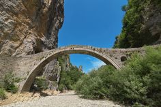 an old stone bridge over a river in the middle of a rocky mountain area with trees on both sides