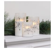 three white candles with snowflakes on them are sitting on a mantle next to pine branches