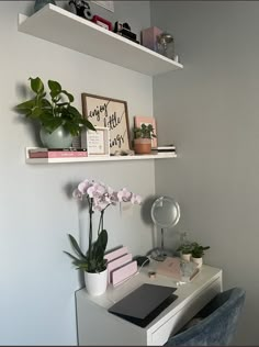 a white desk topped with books and flowers next to a wall mounted shelf filled with plants