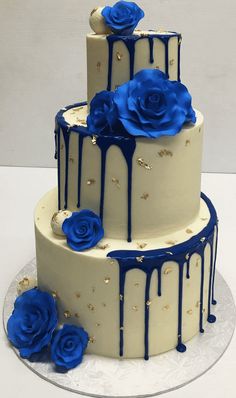 a three tiered cake with blue roses on top and white icing dripping down the side