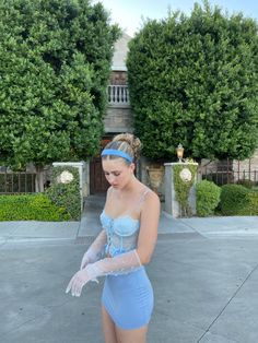 a woman in a blue dress and white gloves is standing on the sidewalk with her hands behind her back