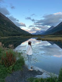 a woman standing on the edge of a body of water with mountains in the background