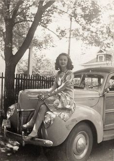 a woman sitting on the hood of an old car in front of a tree and fence