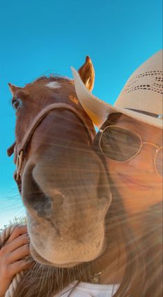 a woman wearing sunglasses and a cowboy hat is hugging a horse's face with the sun shining on her