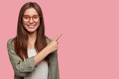 a woman in glasses pointing at something with her finger on the pink background, wearing a green jacket and white t - shirt