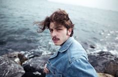 a young man standing on top of a rocky beach next to the ocean with his hair blowing in the wind