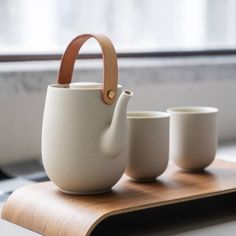 three white cups and a teapot on a wooden tray in front of a window