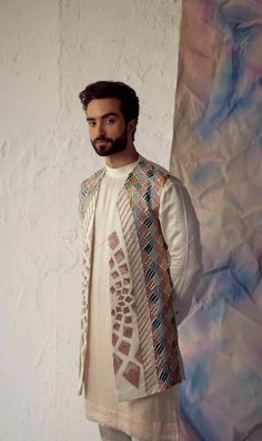 a man standing in front of a wall wearing a white shirt and vest with colorful designs on it