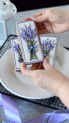 two hands are holding small tiles with lavender flowers on them and ribbons around the edges