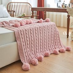 a pink knitted blanket with pom poms on the bottom and sides sitting on a bed