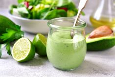 a green smoothie in a glass jar with a spoon next to it, surrounded by limes and avocado