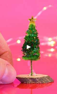 a tiny christmas tree is being held by a finger on a pink surface with lights in the background