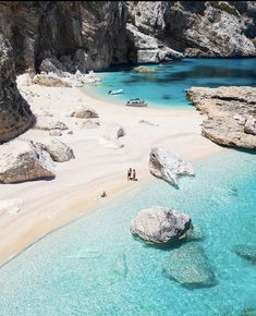 two people are walking on the beach next to some rocks and clear blue ocean water