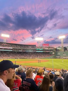 people sitting in the stands watching a baseball game at sunset or dawn with pink and purple clouds