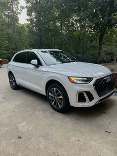 a white audi suv parked in a parking lot next to some trees and bushes on the other side of the road