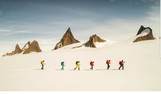 a group of people walking across a snow covered field next to tall mountain peaks in the distance