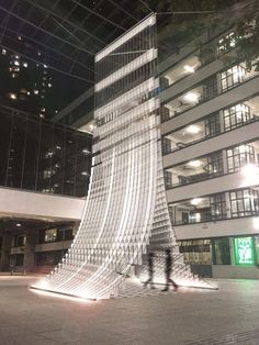 two people walking in front of a tall building at night with lights shining on it