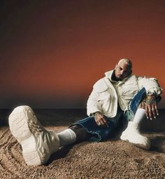 a man sitting on the ground with his feet up and wearing white shoes, in front of an orange sky