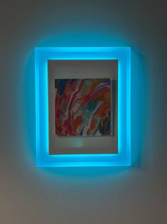 an abstract painting is framed in a white frame on the wall with blue light coming from it