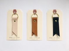 three leather keychains with tags attached to them on a white surface, one is black, the other is tan