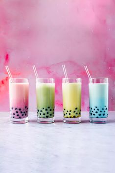 three glasses filled with different colored drinks on top of a white table next to a pink wall