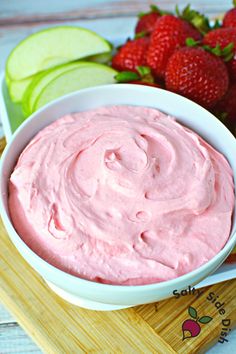 a white bowl filled with pink fruit dip next to sliced apples and strawberries on a cutting board