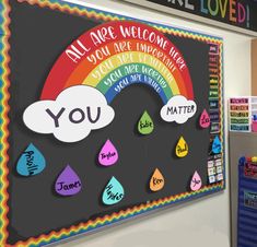 a classroom bulletin board with rainbows and raindrops in the middle, next to a chalkboard that says you are welcome