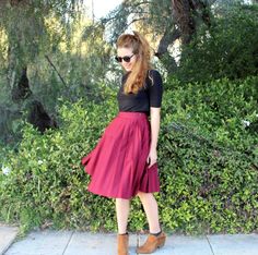 a woman standing on the sidewalk wearing a red skirt and black shirt with brown boots