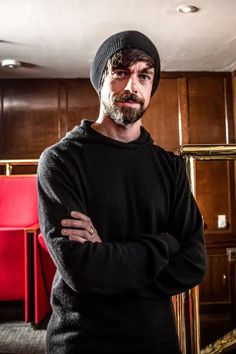 a man with his arms crossed wearing a black sweater and beanie standing in front of wooden cabinets