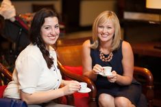 two women sitting on chairs holding cups and saucers in their hands, smiling at the camera