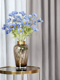 a vase with blue flowers sitting on top of a table next to a white curtain