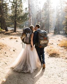 a bride and groom walking through the woods with backpacks on their backs, kissing