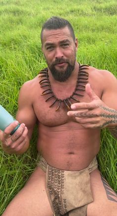 a shirtless man sitting in the grass holding a hair dryer and looking at the camera