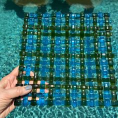 a hand holding a blue and green mosaic tile next to a swimming pool with clear water