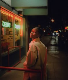 a woman standing on the sidewalk in front of a store at night with neon lights