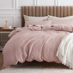 a bed with pink comforter and pillows in a white room next to a table